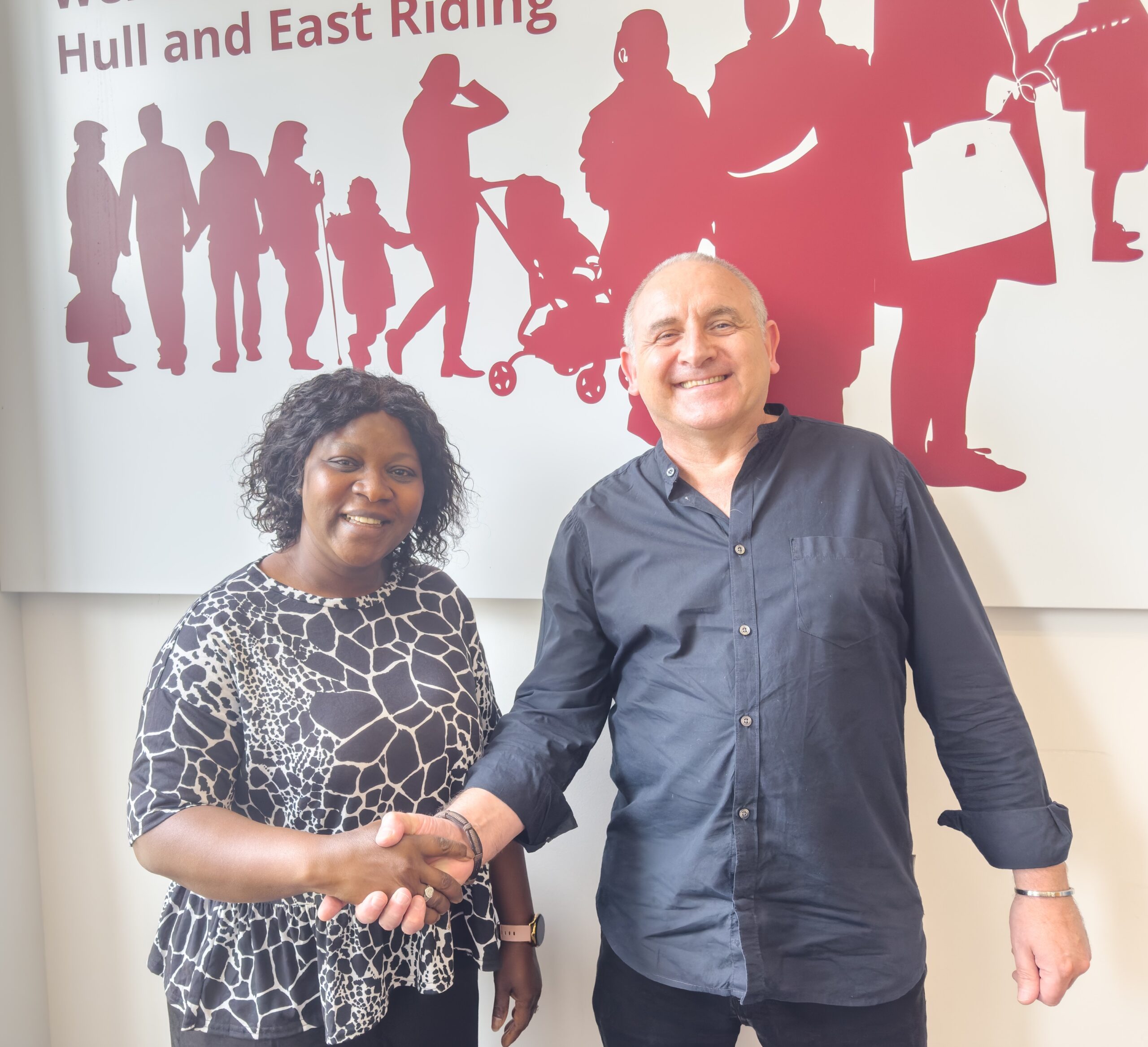 Photograph shows Binta shaking hands with Jon Wilson in front of a Citizens Advice wall decoration at The Wilson Centre in Hull